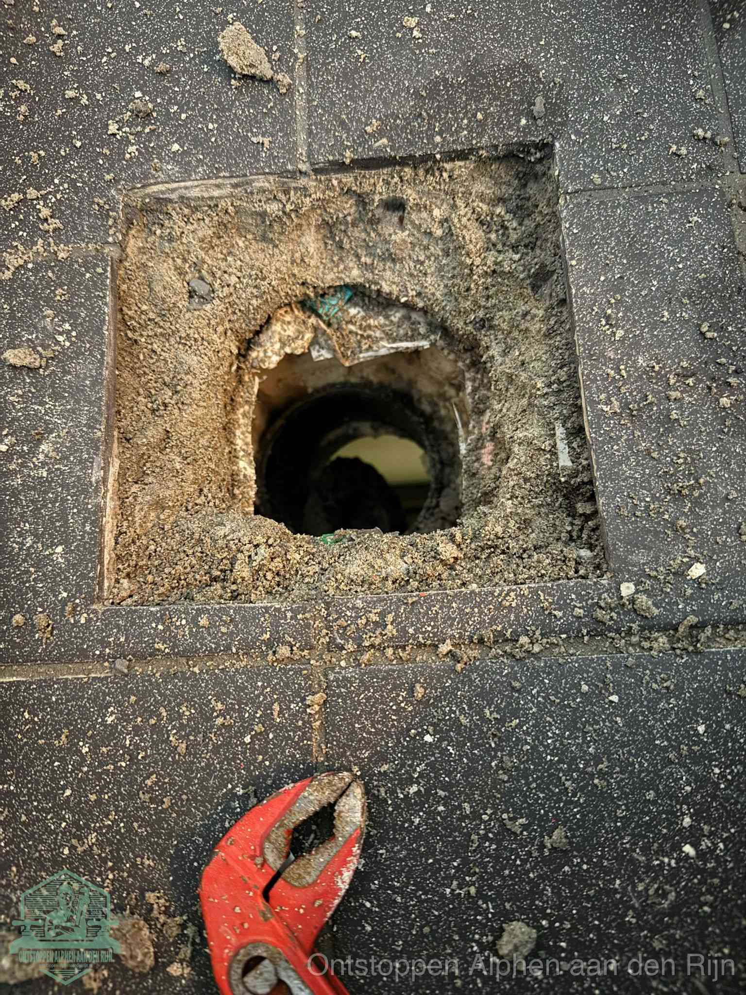 Een geopend rioolputje in een betontegelvloer met losgemaakt rooster en een zichtbare ronde afvoerbuis, het achterhalen waar de afvoerverstopping zit in Alphen aan den Rijn