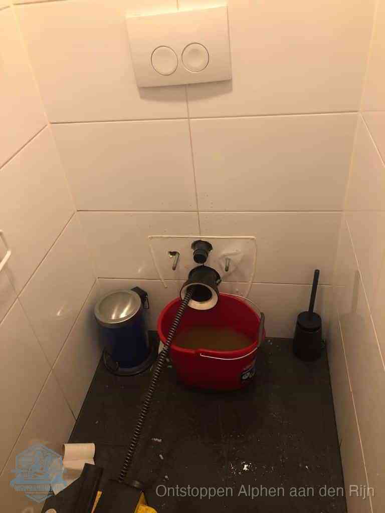 Een ontstopte wand-wc met verwijderde toiletpot waar een ontstoppingsveer in de wanduitgang verdwijnt, het achterhalen waar de verstopping zit in Alphen aan den Rijn