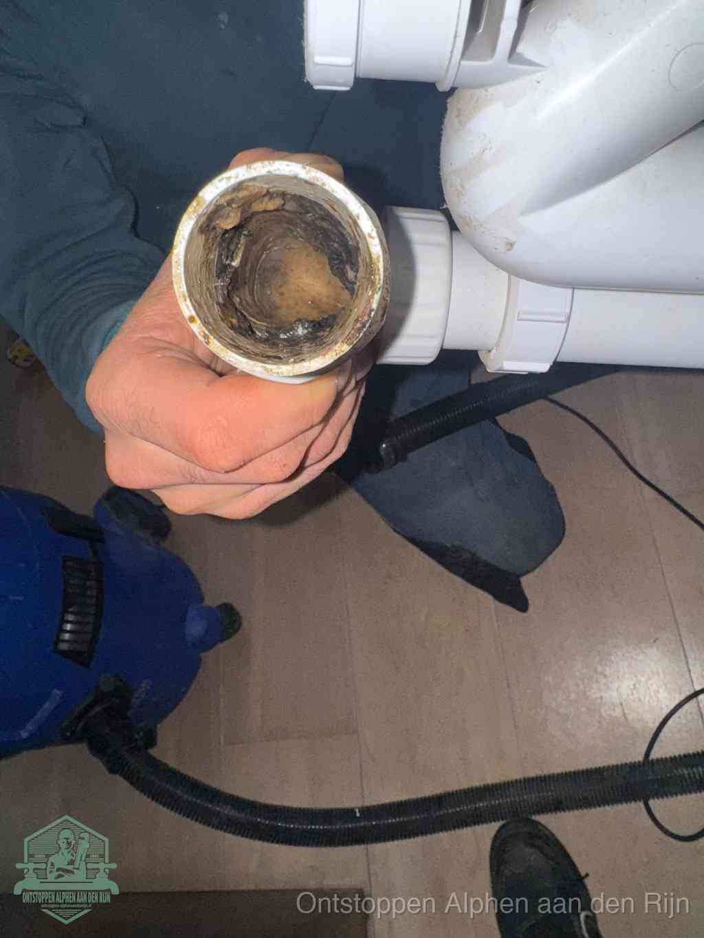Een hand houdt een losgekoppeld PVC-afvoerbuisstuk vast dat van binnen volledig is dichtgeslibd met vet, vuil en aanslag, de meest voorkomende oorzaak van een verstopte afvoer in Alphen aan den Rijn