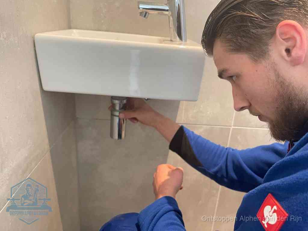 Een monteur draait met de hand de zwanenhalssifon onder een fonteinwasbak in de toiletruimte los om de bron van rioollucht in Alphen aan den Rijn te vinden