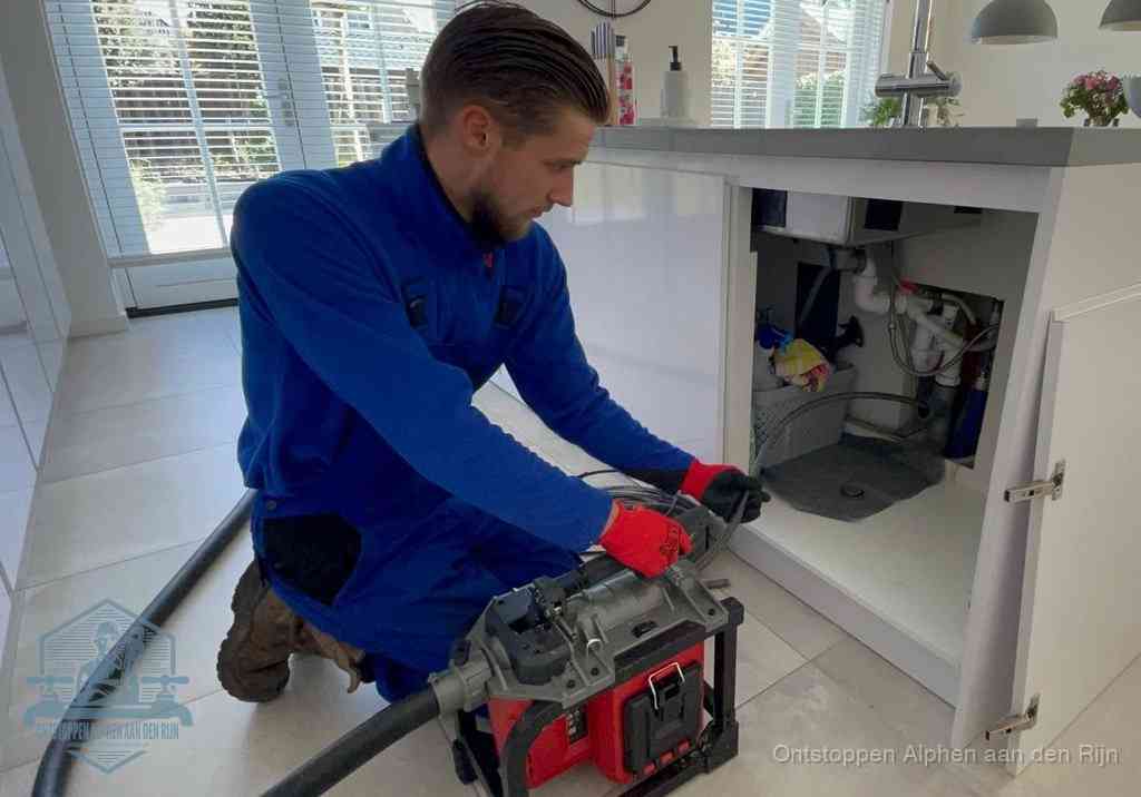 Een monteur voert een professionele elektrische ontstoppingsmachine in de afvoer onder de spoelbak tijdens het gootsteen ontstoppen in Alphen aan den Rijn