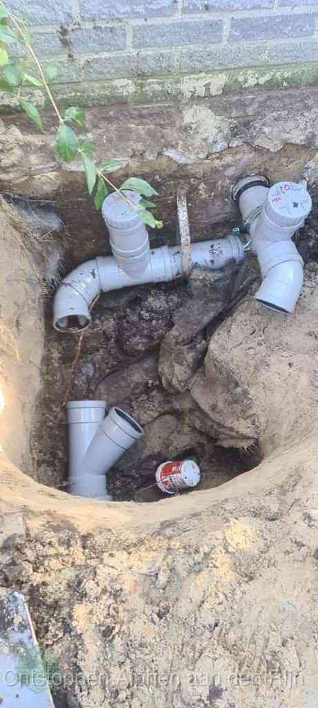 Een opgegraven bouwput langs een huismuur met grijze PVC rioolbuizen die op het hoofdriool worden aangesloten, typisch leidingwerk per wijk in Alphen aan den Rijn